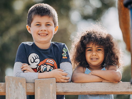 Zwei Kinder lehnen lachend an einem Holzzaun auf dem Spielplatz